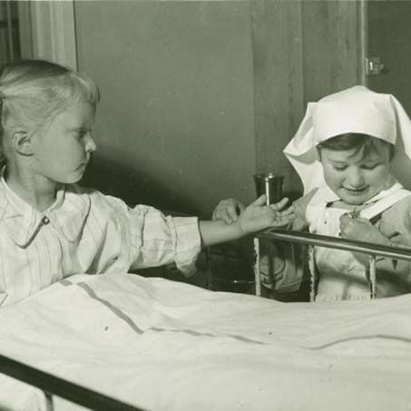 Et barn sitter i en seng og tas pulsen på av et annet barn som er kledd ut som en sykepleier. Historisk foto.