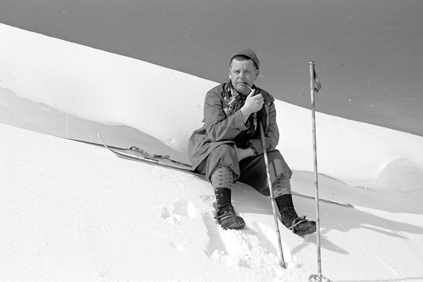En mann med ski og staver, sitter i sola i en snøskavl og røyker pipe. Foto.