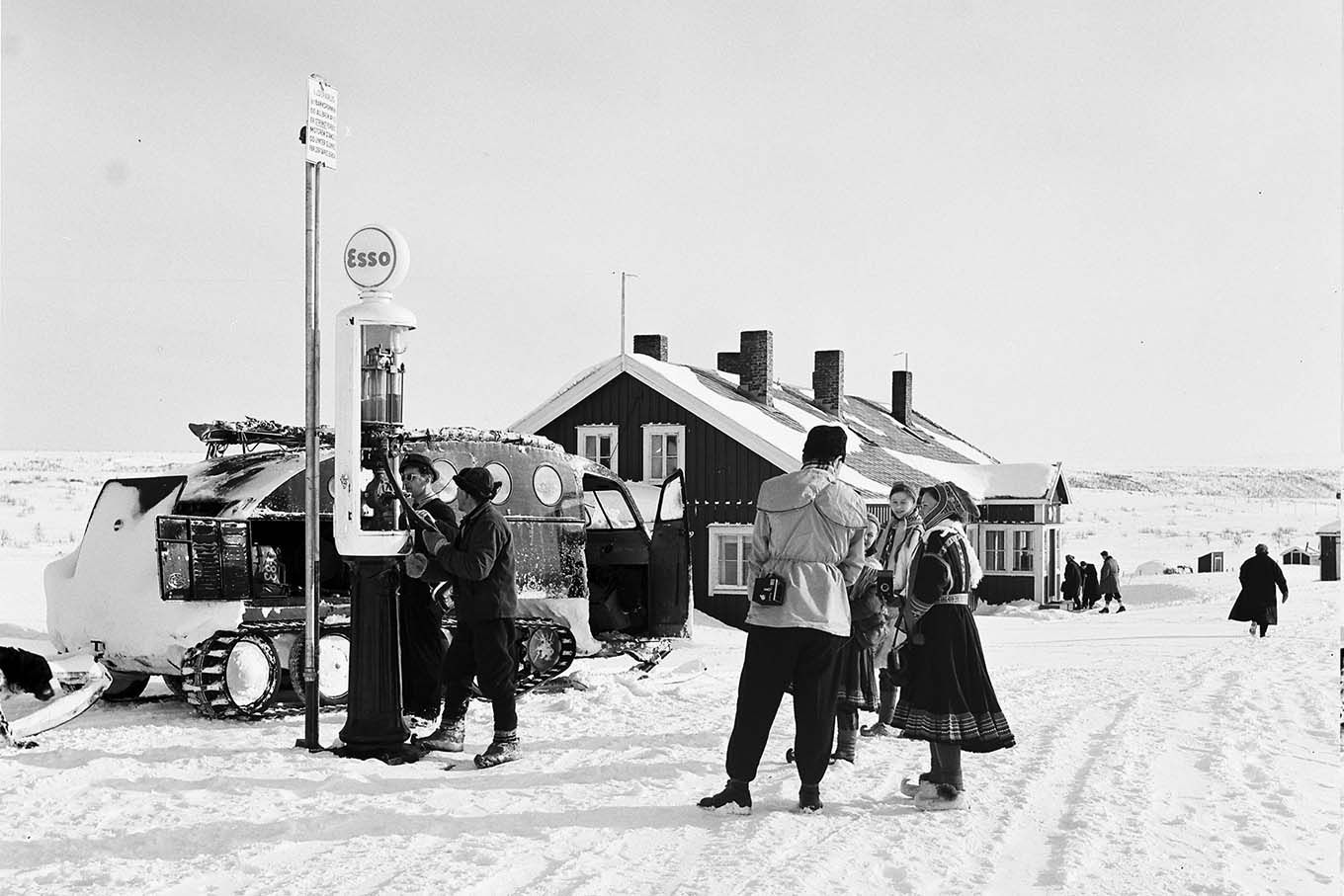 1955. En campingvogn med beltehjul på en bensinstasjon i snødekket landskap. En mann med kamera over skulderen snakker med damer iført samedrakt. Historisk foto. 