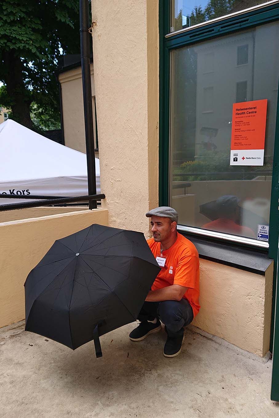 En frivillig og papirløs sitter utenfor helsesenteret med en paraply. Foto.