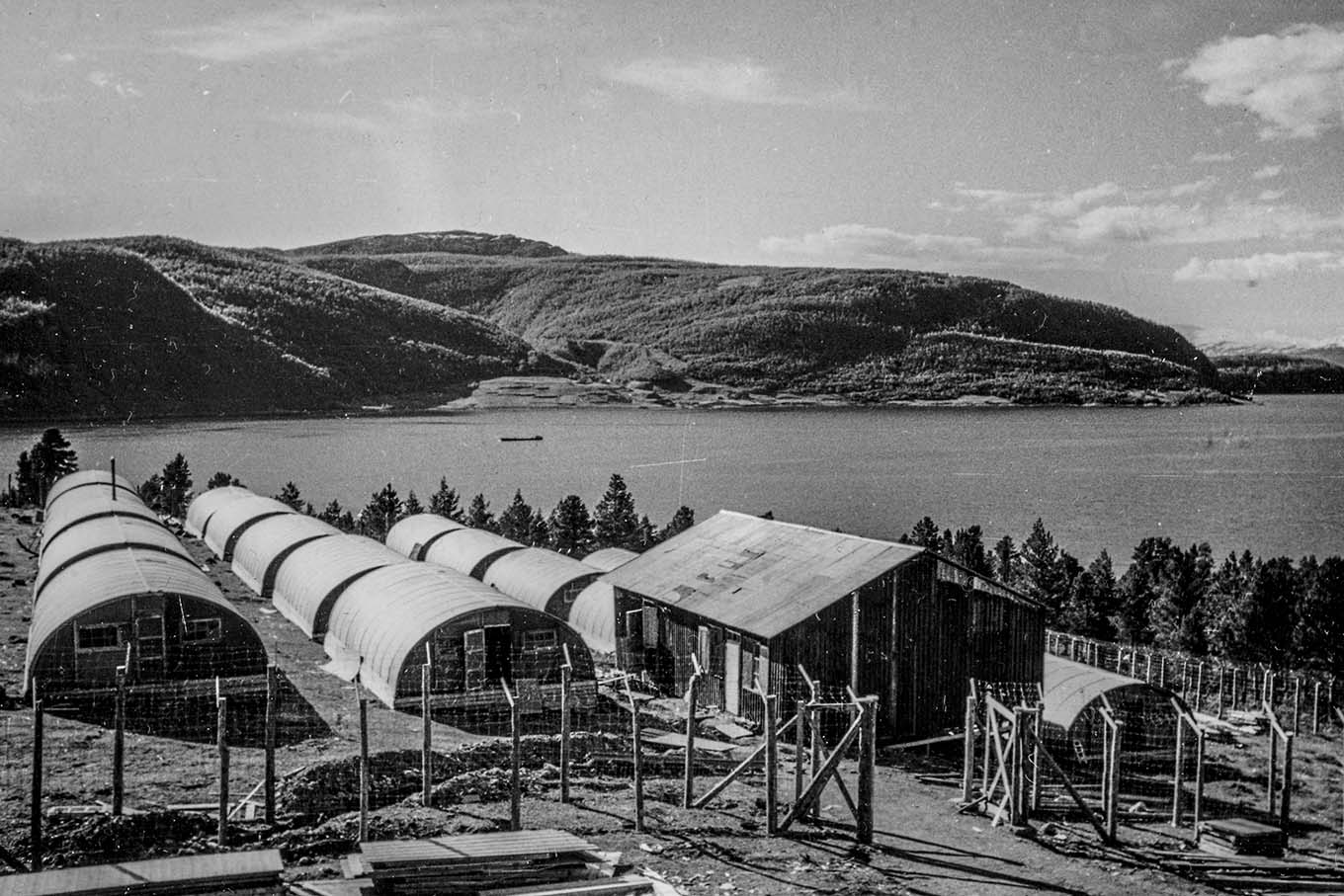 Inngjerdede militærbrakker, i bakgrunnen et vann og fjellandskap. Foto.