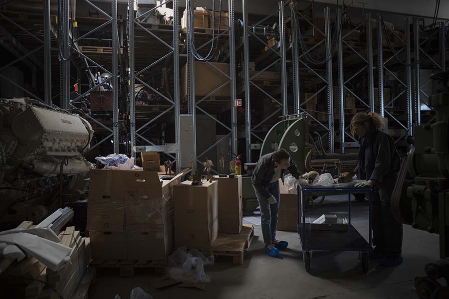 To mennesker står og studerer menneskeknokler i et stort lagerlokale. Det er flere store gjenstander på gulvet og på høye hyller. Foto.