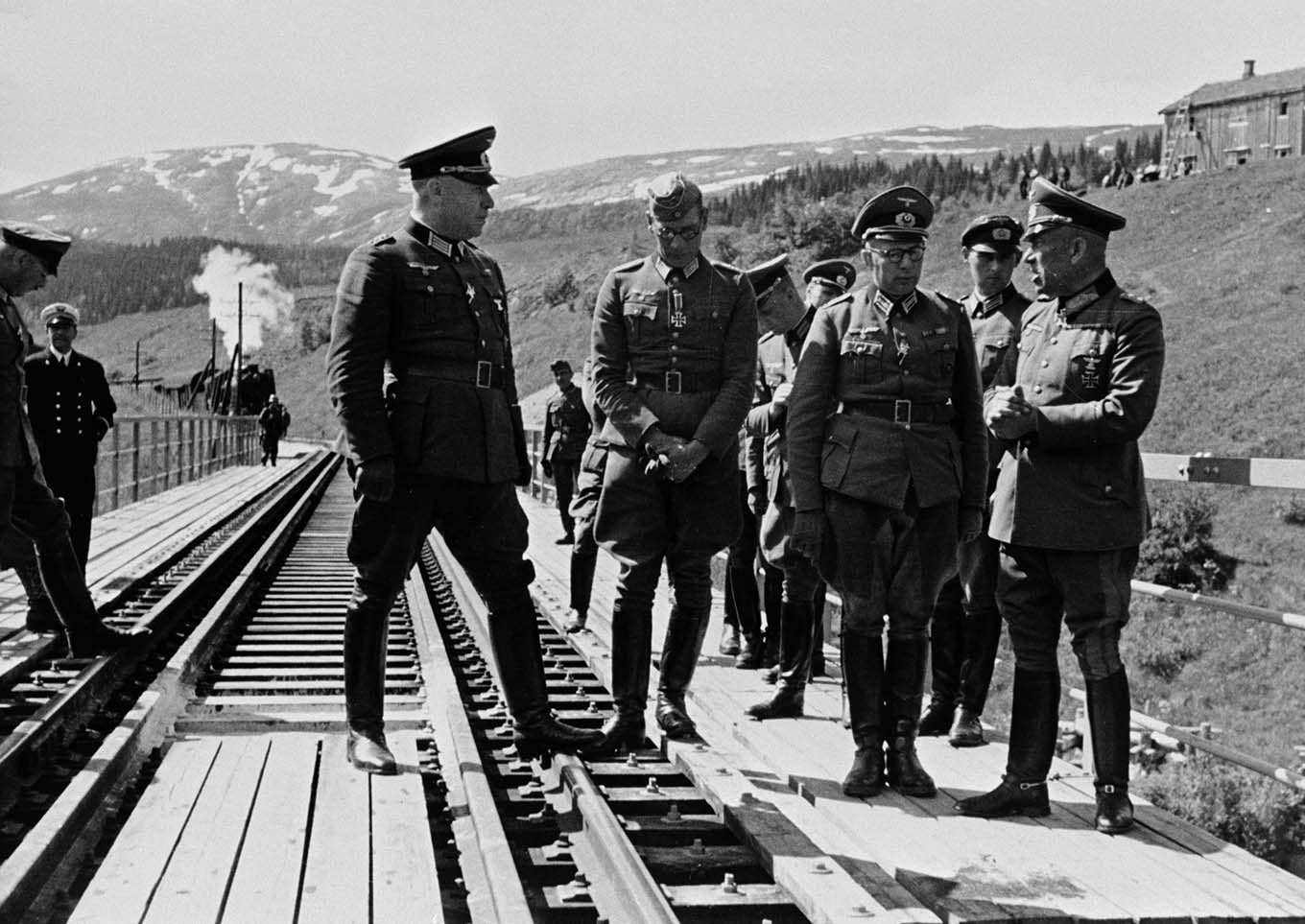 Men in military uniform wait for the train steaming into the station in the background. Photo. 