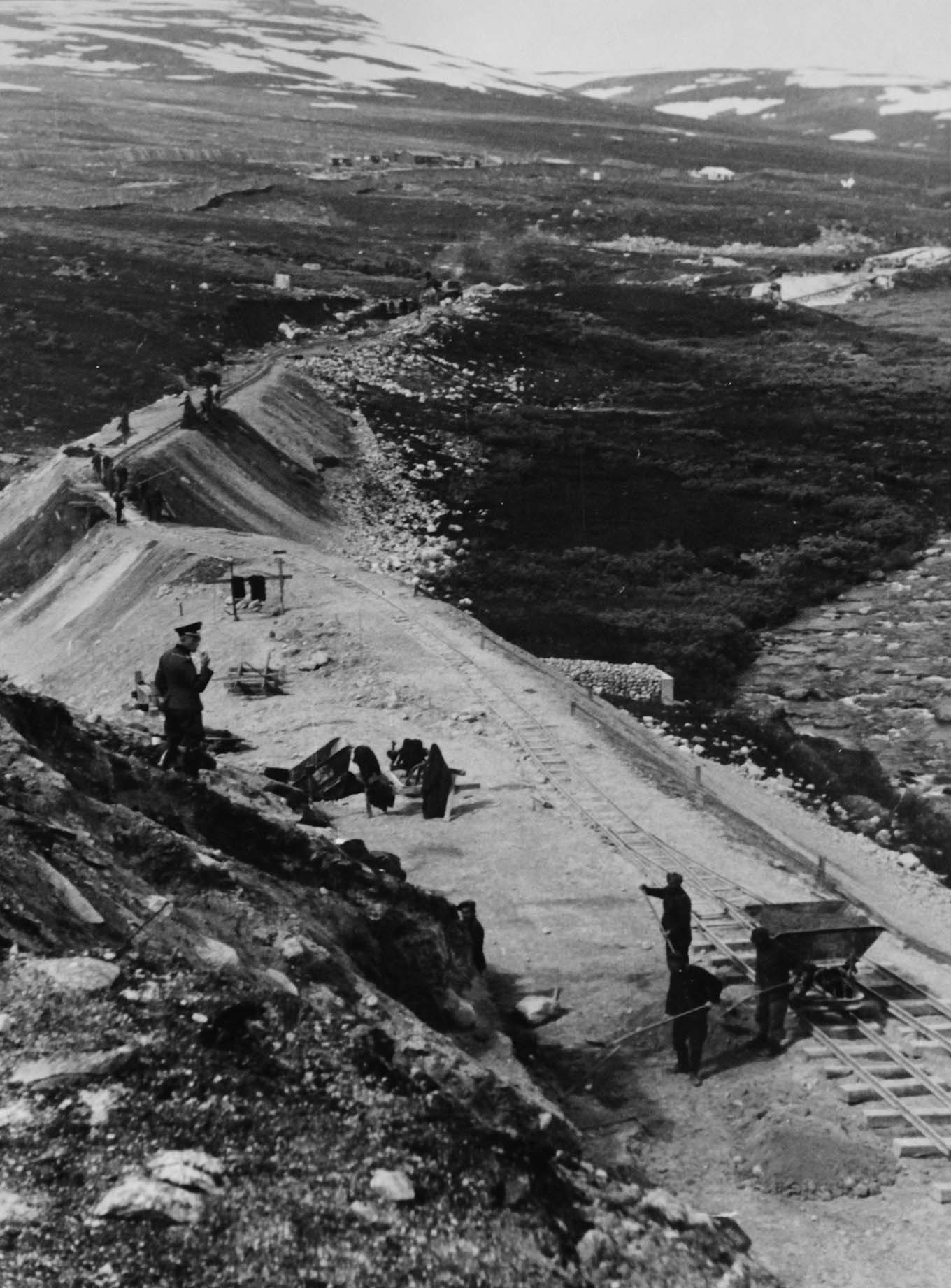 A soldier guards prisoners of war at work by the railway track through a mountain landscape. Photo. 