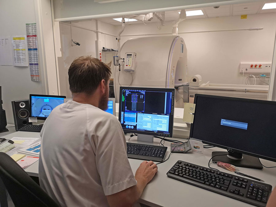 En mann sitter foran flere dataskjermer og administrerer en stor medisinsk skanner.