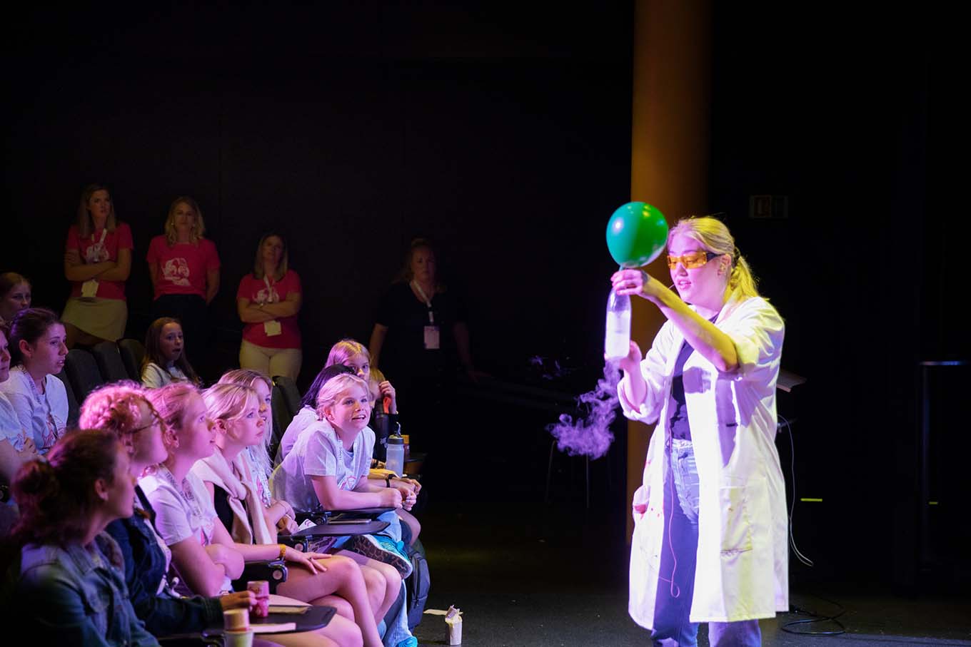Woman in lab coat demonstrates experiment with balloon and smoke in front of audience. Photo.