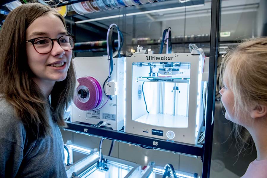 To jenter står ved en 3D-printer som er i gang med printing. Foto.