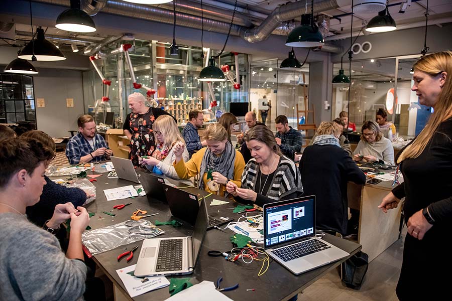 Mange voksne mennesker sitter på langbord og bygger elektronikk. Foto.