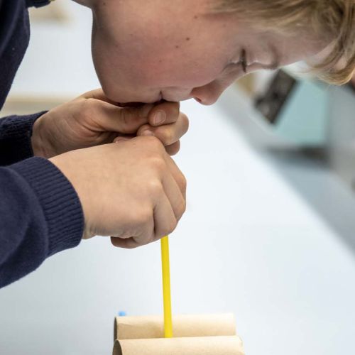 En gutt blåser i et sugerør over to pappruller. Foto