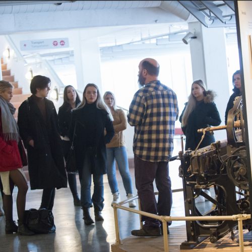 Man at a mechanical loom chatting in front of eight young adults. Photo
