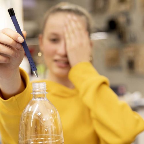 Jente holder venstre hånd foran venstre øye mens hun holder en penn i høyre hånd og fører pennen ned i en plastflaske. Foto