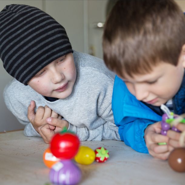 Two boys lean over a table with various wooden figures in different colors representing fruits, berries and vegetables. Photo