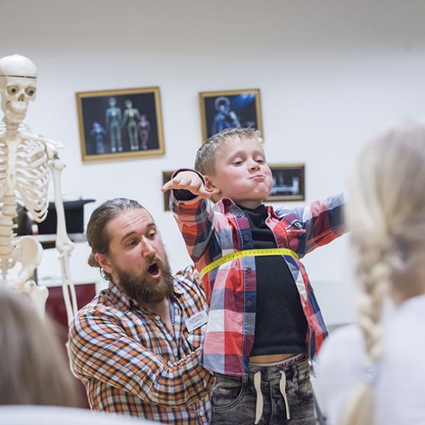 Man measures the chest of a child who holds his arms out and makes a face, in a classroom with skeletons. Photo