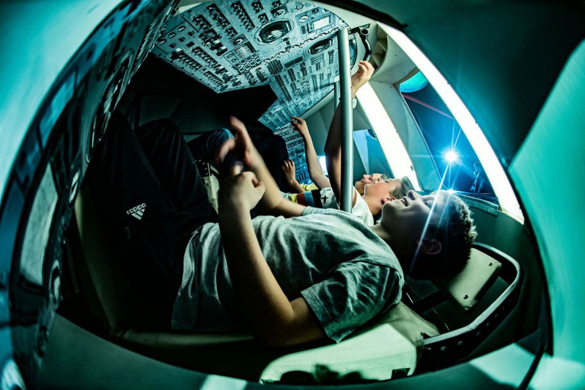 Tre ungdommer sitter inni i en modell av en romkapsel. Foto.