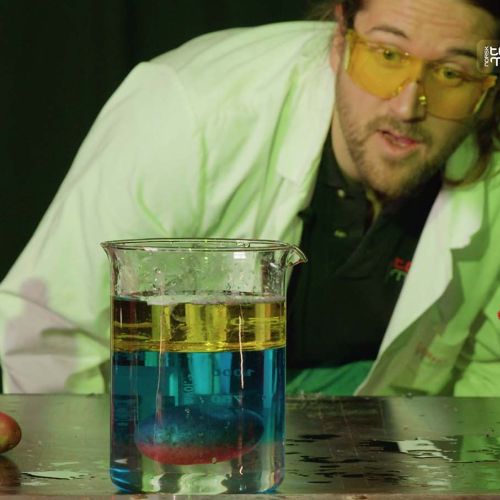 Mann med verneutstyr ser på et begerglass, hvor det flyter en potet i midten av tre væsker i forskjellige farger. Foto