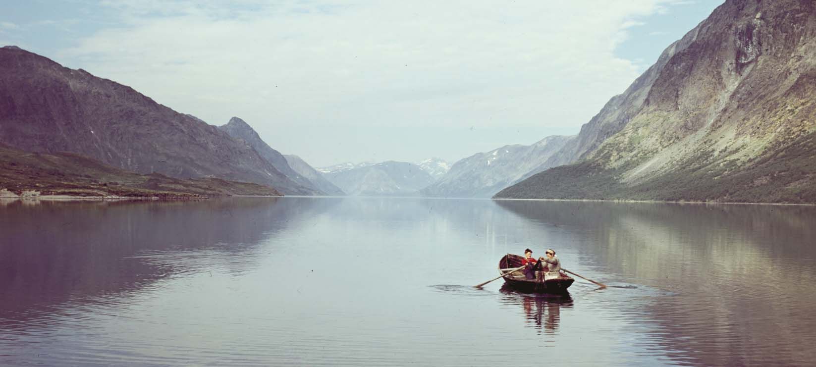 To personer ror på et vann mellom to fjellsider. Minner om maleriet "Brudeferden i Hardanger". Foto.