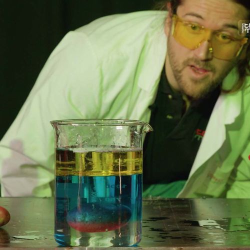 Mann med verneutstyr ser på et begerglass, hvor det flyter en potet i midten av tre væsker i forskjellige farger. Foto