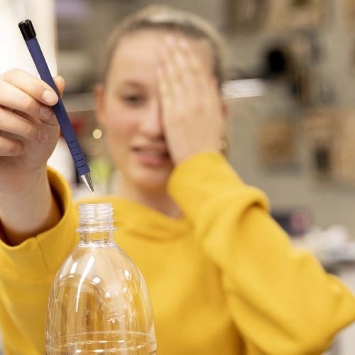 Jente holder venstre hånd foran venstre øye mens hun holder en penn i høyre hånd og fører pennen ned i en plastflaske. Foto