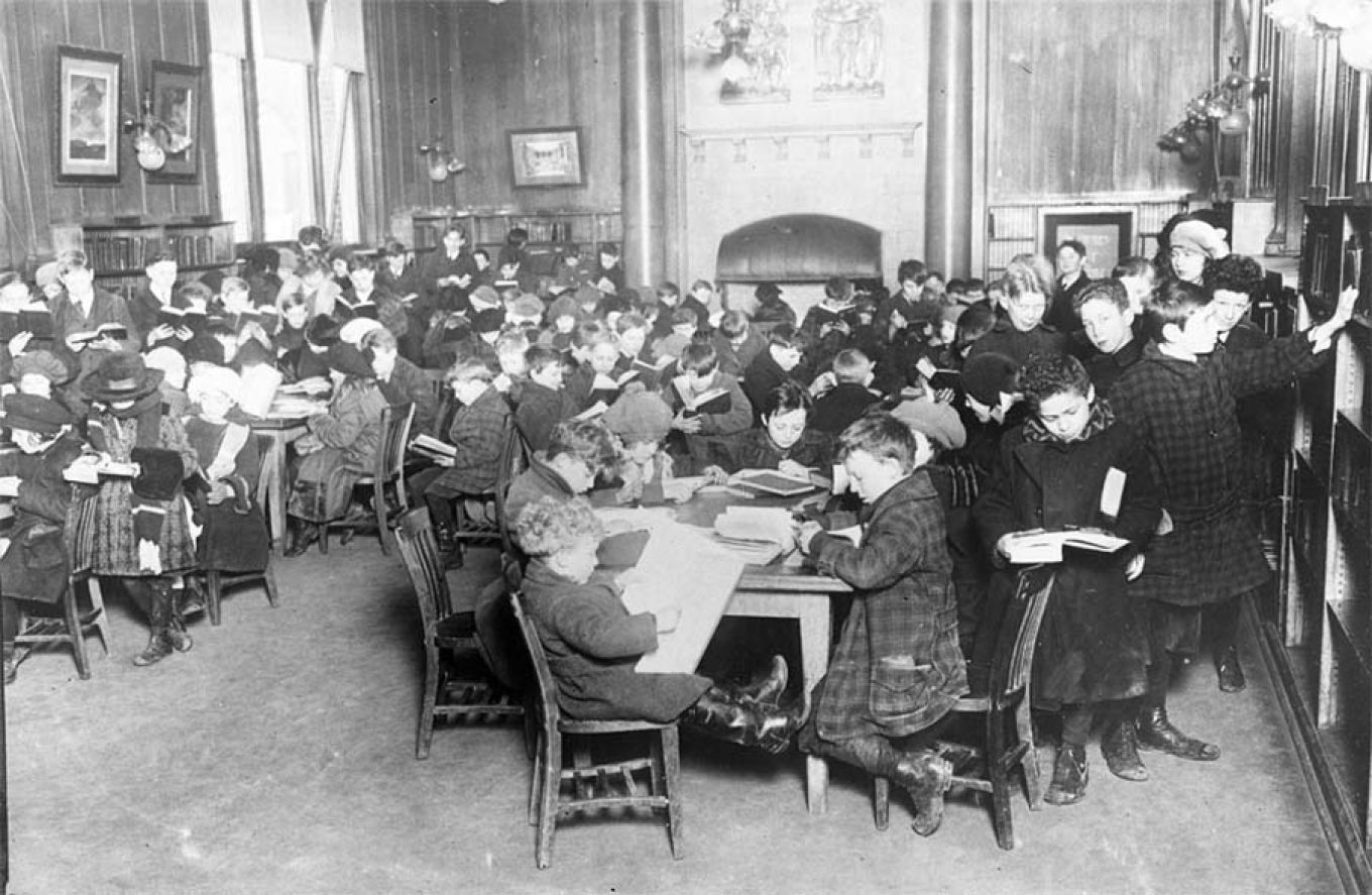 Gammelt bilde av skolebarn som sitter rundt flere bord og leser i bøker. Foto.