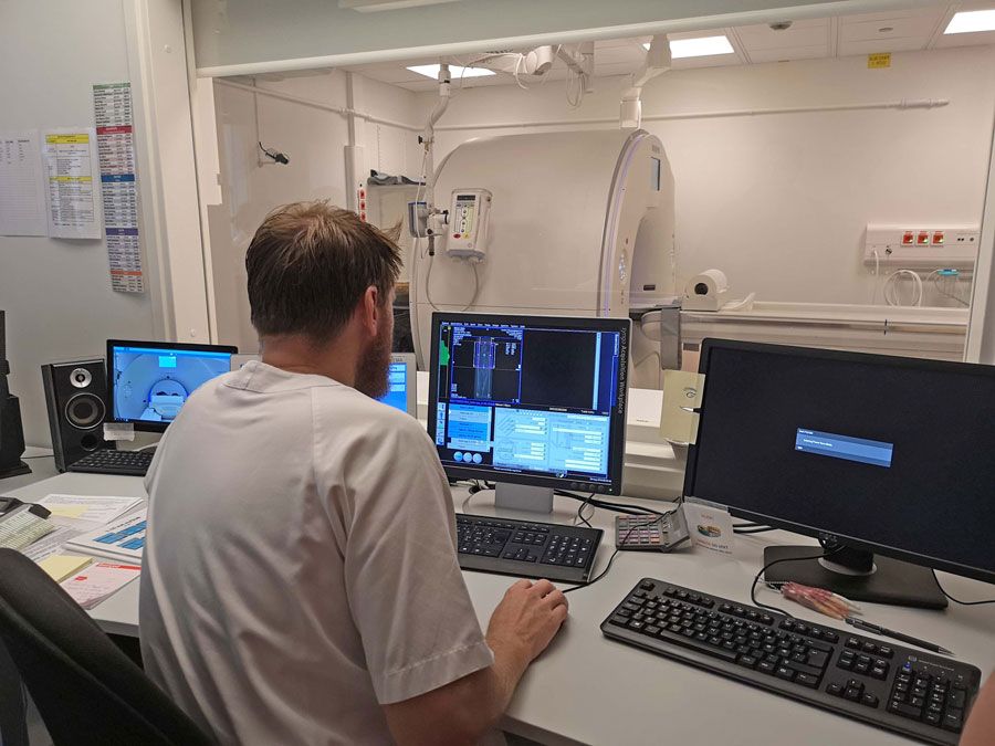 En mann sitter foran flere dataskjermer og administrerer en stor medisinsk skanner.