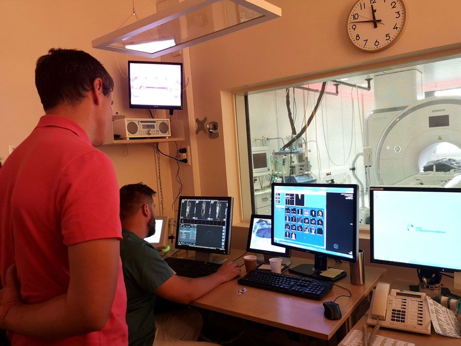 To menn står ved flere dataskjermer og kontrollerer en stor medisinsk skanner.