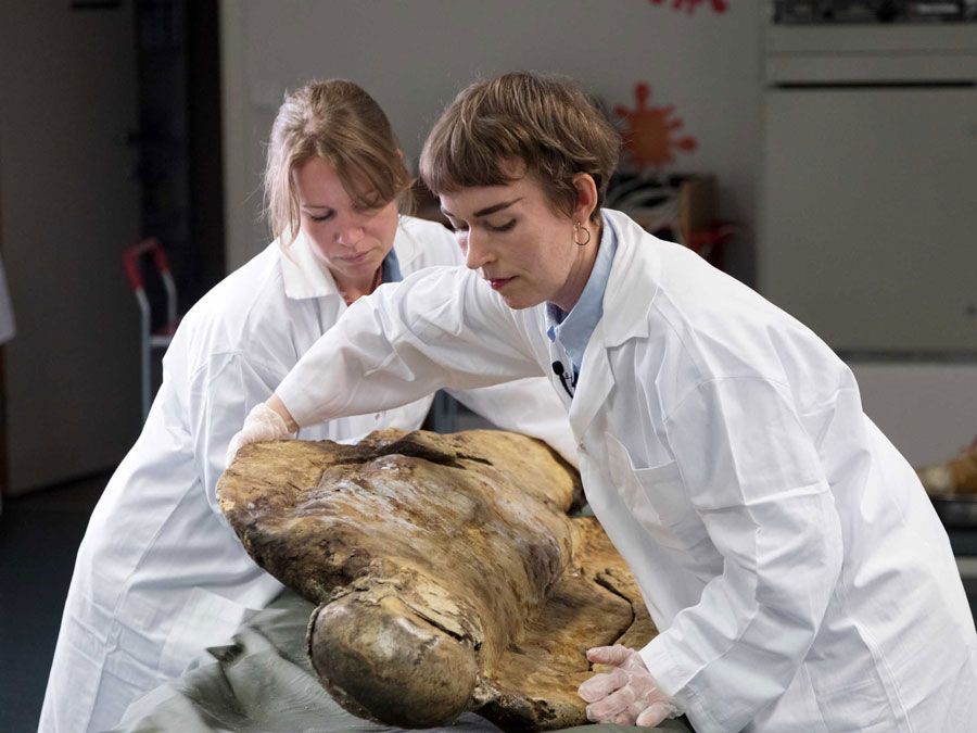 To kvinner i labfrakk hjelper hverandre å snu et gammelt kolera lik.