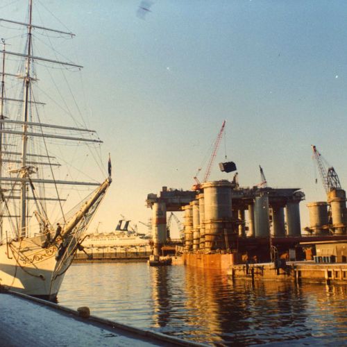 Hvit seilbåt ligger til havn sammen med en flytende oljerigg. Historisk foto.