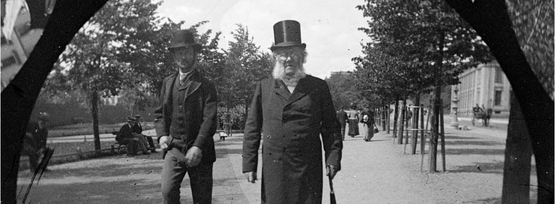 Knappehullsfoto av Henrik Ibsen i flosshatt, og en annen mann, som spaserer langs Karl Johans gate. Foto.