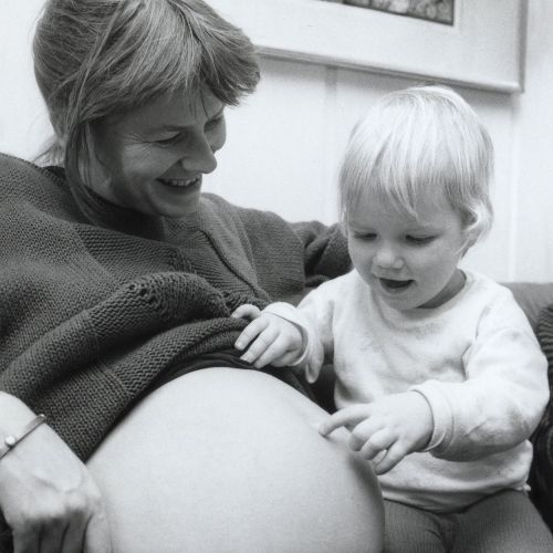 Sort-hvitt bilde av en smilende gravid mor som holder et ungt barn. Perfekt illustrasjon for Fødselskafé på Teknisk museum under morsdagen, der foreldre og besteforeldre deler erfaringer om fødsel for å forbedre fødselskulturen i Norge.