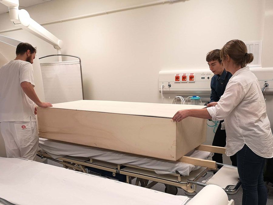 To kvinner og en mann står i et rom med medisinsk utstyr. En kiste ligger på en båre.