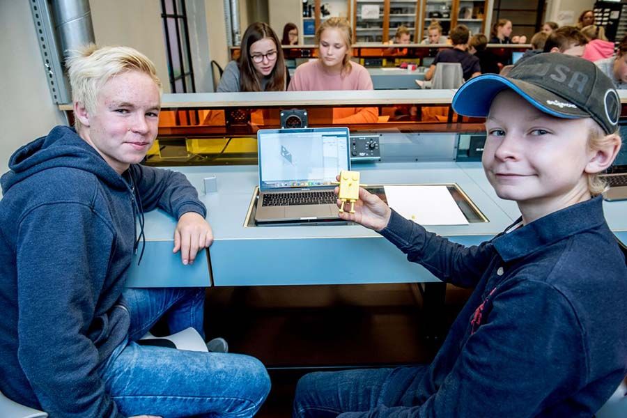 To gutter sitter ved et bord med en datamaskin og viser frem en 3D-printet figur. Foto.